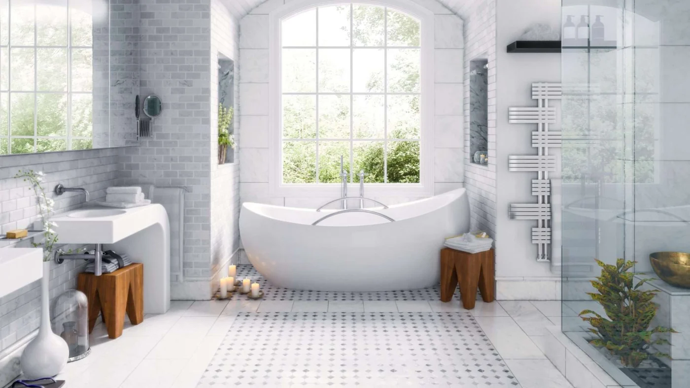 All white bathroom with stand-alone tub, showers, and individual vanities. White brick walls with a window overlooking trees. Candles on the floor and plants in the shower.