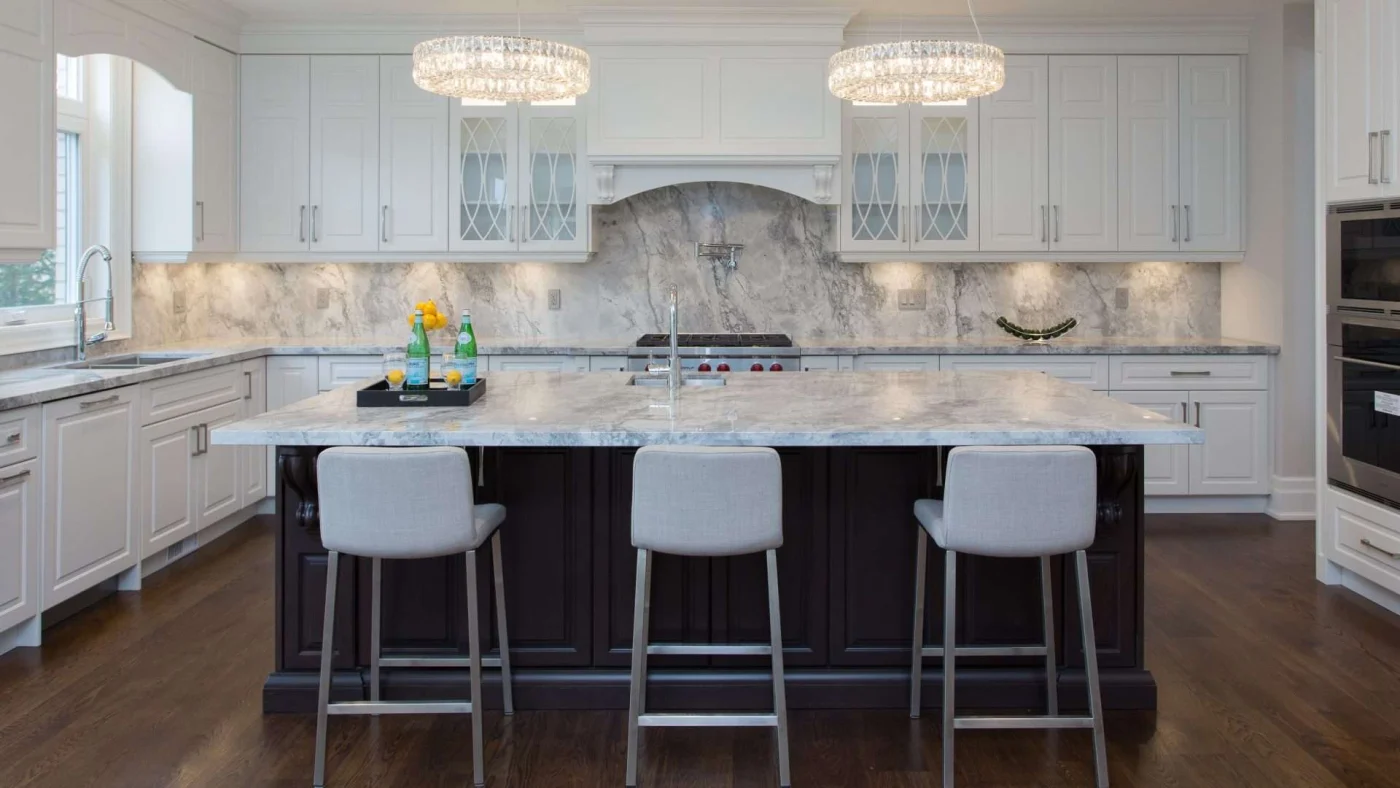 A kitchen with marble countertops and backsplash, an island with seating for three, white cabinets, and stainless steel appliances
