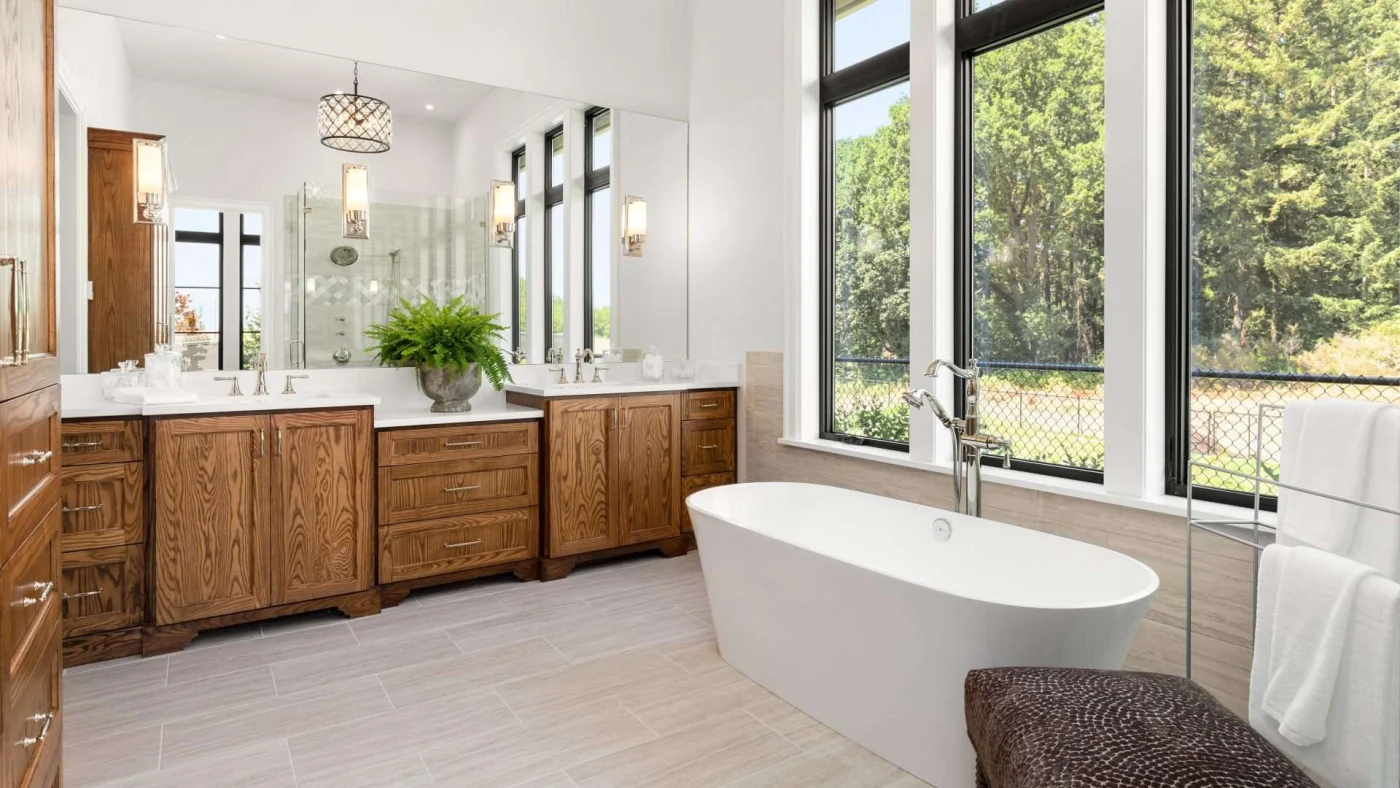 A stylish bathroom with a soaker tub beneath the windows, a brown wood vanity, and a large wall-to-wall mirror with a shower in the reflection
