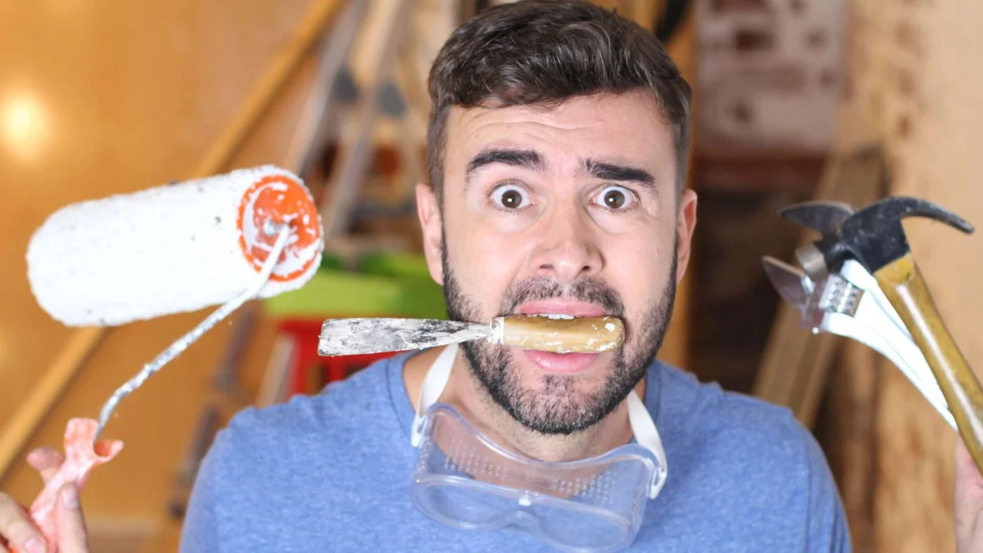 A frazzled man stands with a chisel in his mouth, a hammer in one hand, and a paint roller in the other
