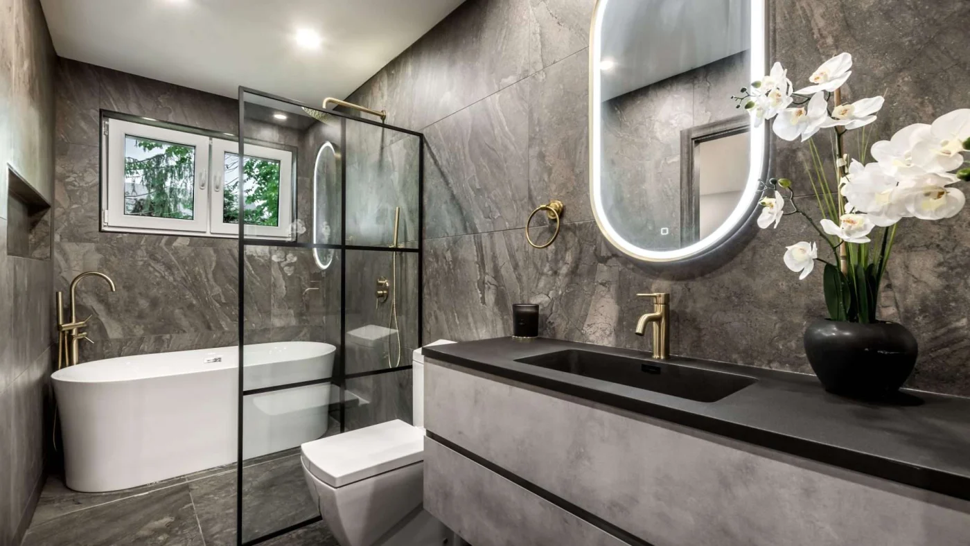 Angled interior view of a beautifully redone bathroom with grey tile and a freestanding tub and shower behind a glass panel