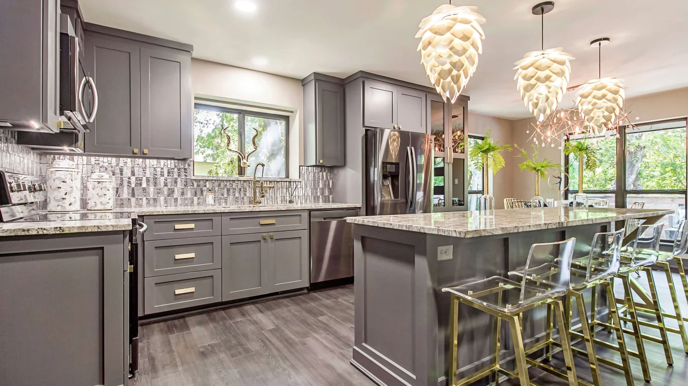 A kitchen remodel with gray shaker style cabinets, light colored granite countertop, matching backsplash, and a large island with clear bar stools.