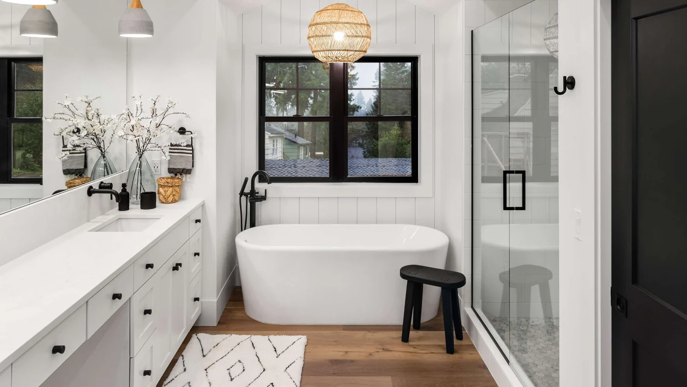 A modern bathroom remodel with white walls, light wood flooring, a freestanding white bathtub, and a glass-enclosed shower.