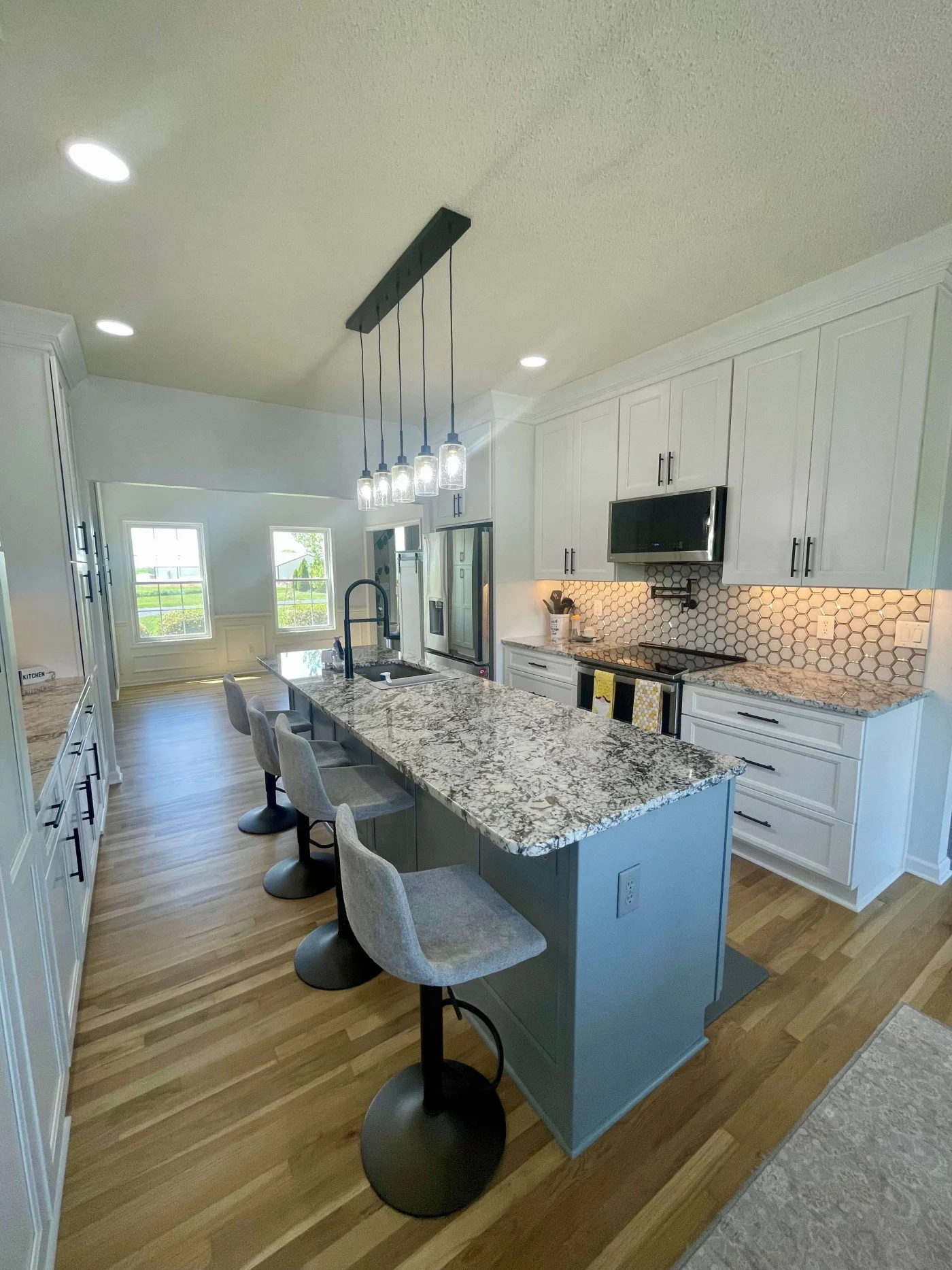 A modern kitchen remodel with a large island featuring a light colored granite countertop, white upper and lower wood cabinets, and a microwave with a patterned tile backsplash.