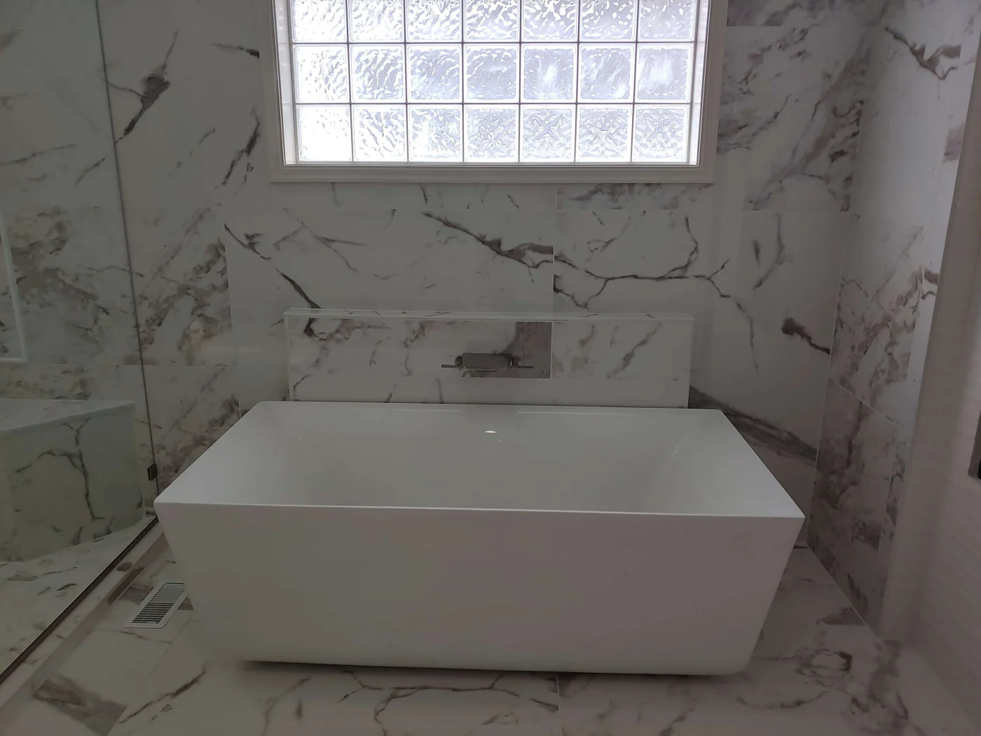 A modern bathroom remodel with a white freestanding rectangular tub, marble-patterned walls and floors, and a horizontal block window.
