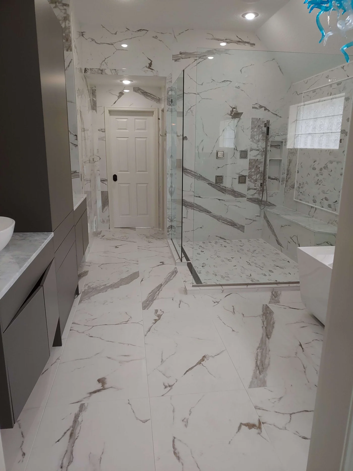 A modern bathroom features white marble-look tiles on the floor and walls, a large glass-enclosed shower, and a freestanding bathtub.