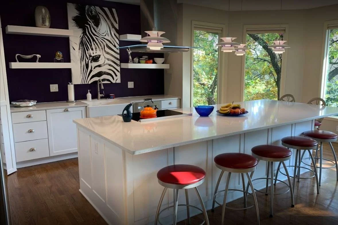 A perspective of a beautifully remodeled kitchen in a Kansas City home with a unique solid dark blue backsplash behind the sink with floating shelving and a large painting in the center, modern lighting over the large white island with white stone countertop, and red stools.