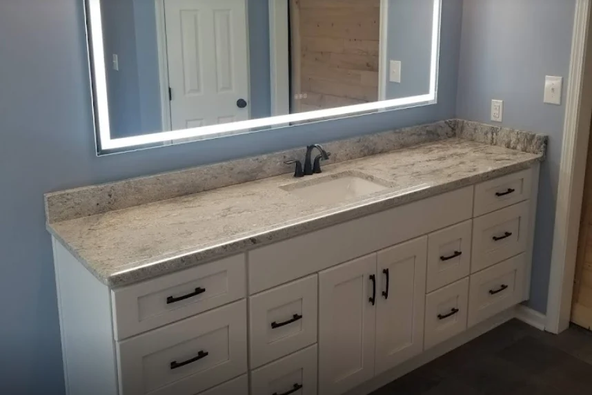 A beautiful new vanity in a Kansas City bathroom after a remodel with Jericho Home Improvements, with stone countertops, a single small sink, dark hardware, and white cabinetry and drawers. There’s a large mirror on the wall above it with white lighting in the frame of the mirror.