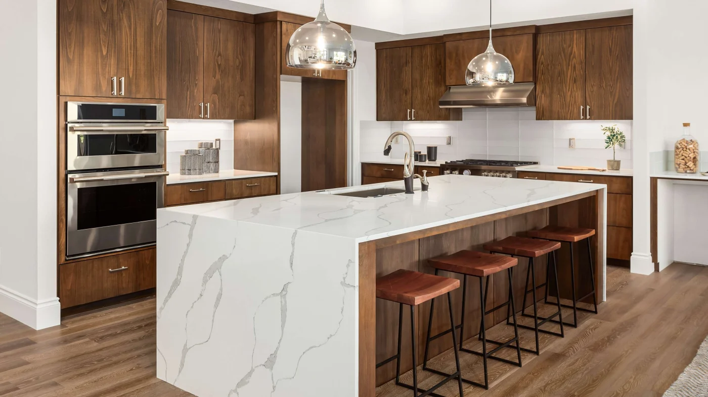 A modern kitchen remodel with dark wood cabinets, a waterfall quartz countertop, stainless steel appliances, light colored wood floors, and four bar stools