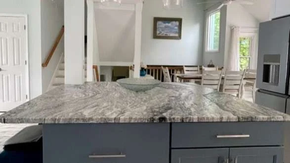 A modern kitchen remodel with a kitchen island with a gray granite countertop and dark gray cabinets with a staircase, dining area, and windows in the background.