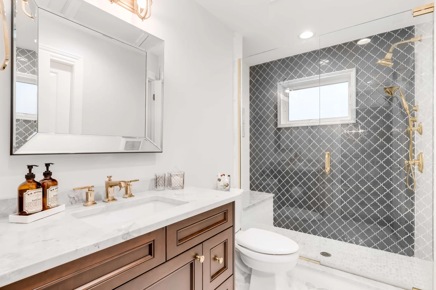 A bathroom with a wooden vanity, marble countertop, white toilet, and a walk-in shower with a glass door and gold fixtures.