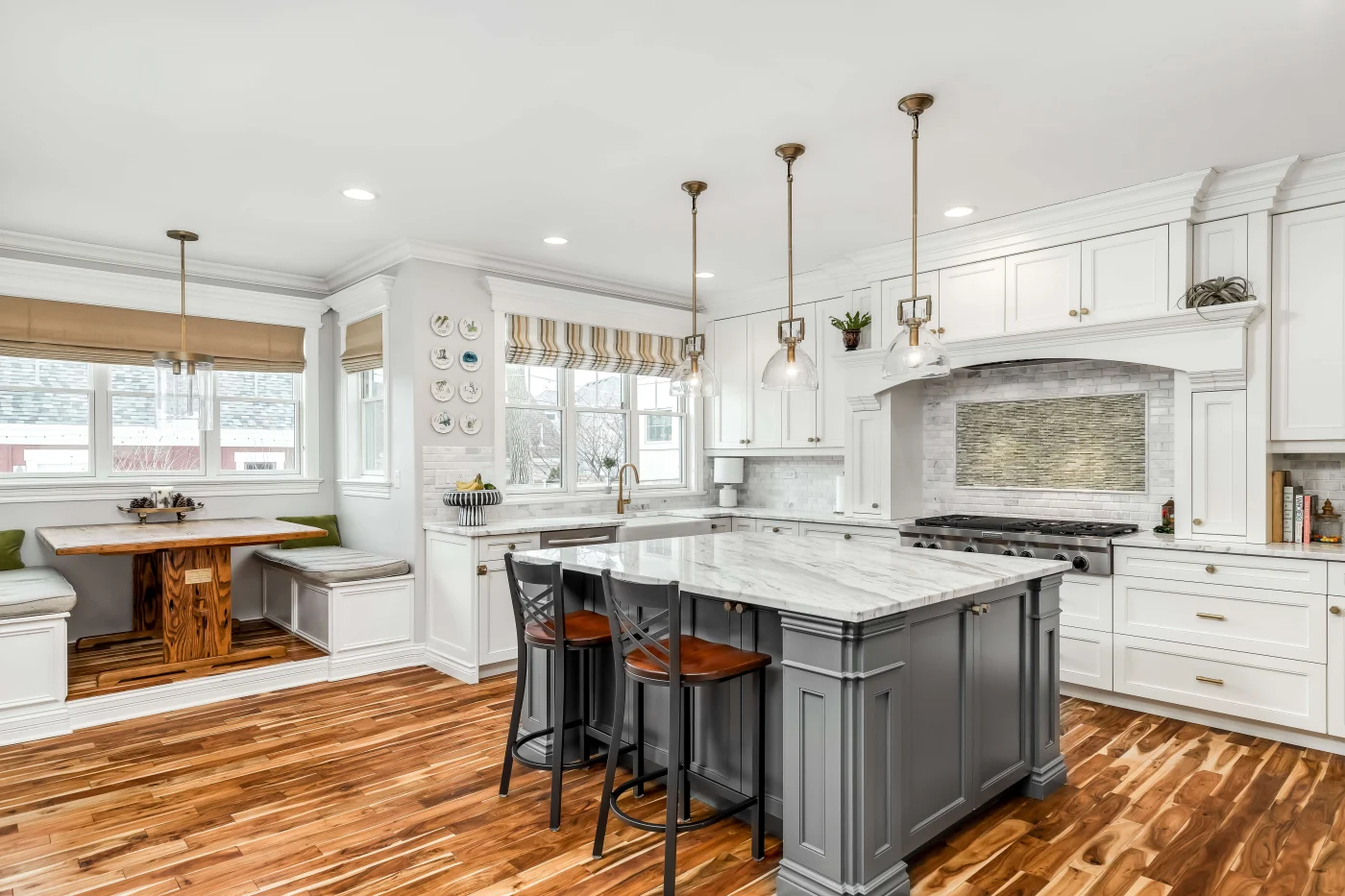 A white kitchen with white cabinets, a gray kitchen island with a marble countertop, and a booth with cushion seats and a wooden table.