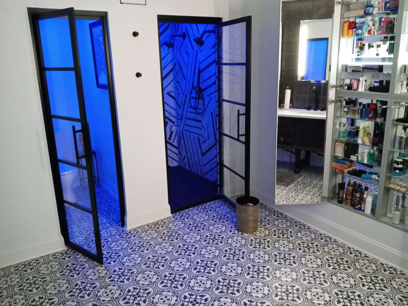 A bathroom with black and white floors, white walls, white toilet in a room with a glass door and purple lighting, and a shower in a room with purple lighting and a glass door.