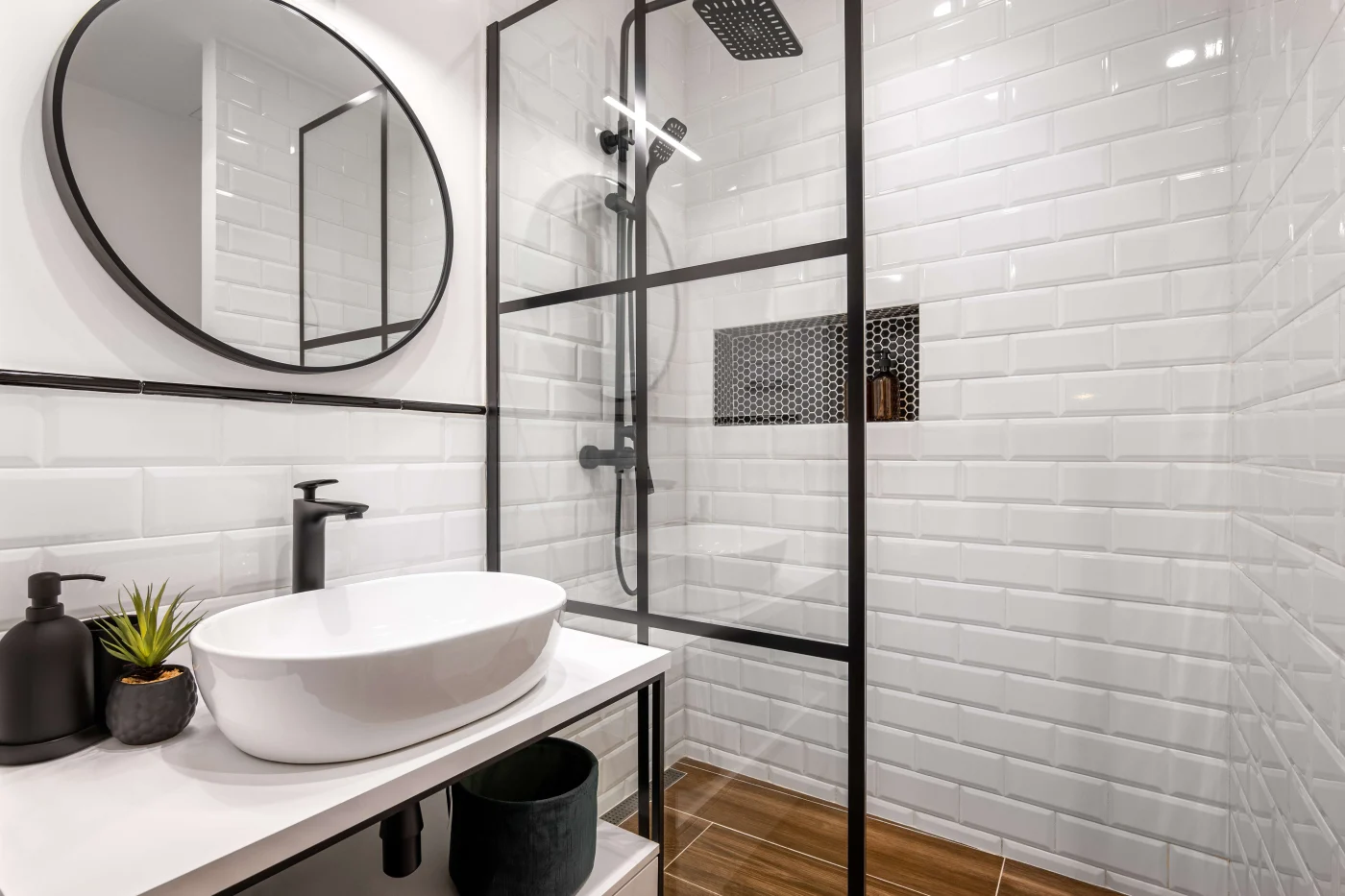 A walk-in shower with a glass wall and black fixtures in a white bathroom with a white sink and a circular mirror above it.