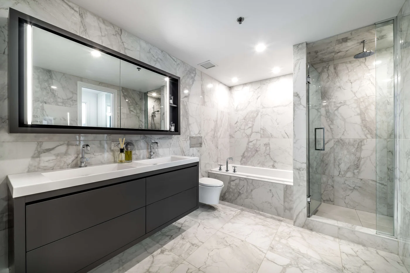 Beautiful Bathroom Remodel In Blue Springs, MO | Jericho A gray and white bathroom with a dark vanity, white sinks, a longer mirror with storage space, a white bathtub, and a walk-in shower with a glass door.