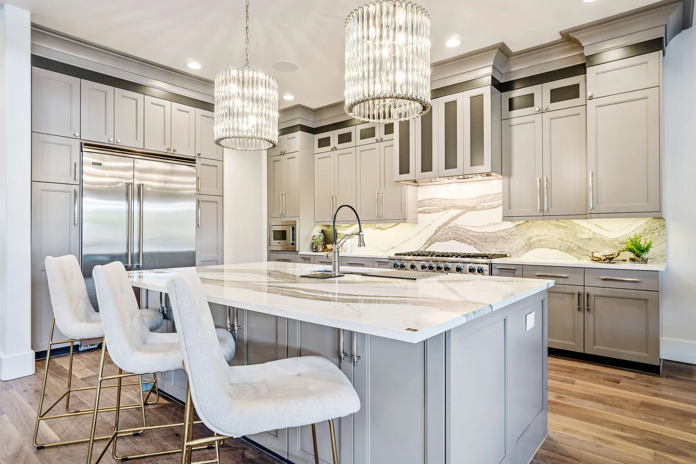 Efficient Kitchen Remodel In Blue Springs, MO | Jericho A kitchen with wooden flooring, gray cabinets, a gray island with a white and gray countertop, white chairs, steel appliances, and circular overhead lights hanging over the island.