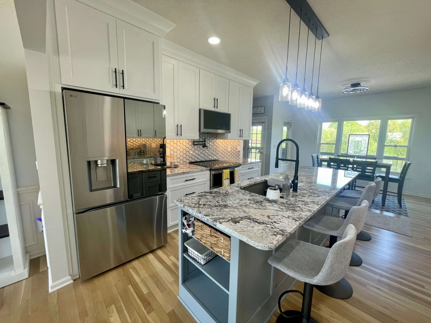 Excellent Kitchen Remodel In Lenexa, KS | Jericho A kitchen with wooden flooring, white cabinets, a blue kitchen island with black and gray countertops, gray chairs, steel appliances, overhead lighting, and a dining room in the background.