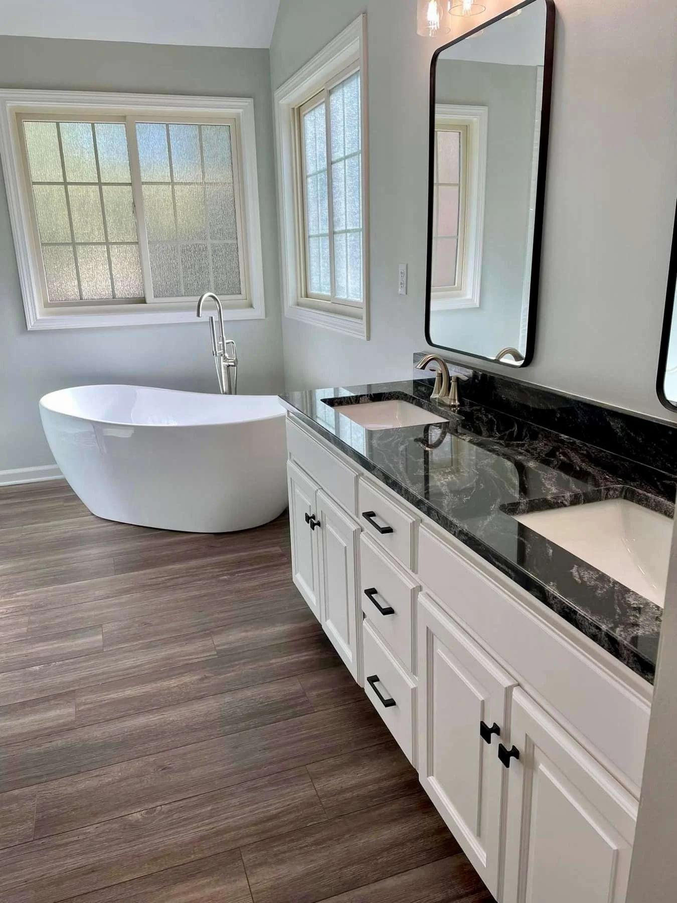 A gray bathroom with wooden flooring, a white freestanding bathtub, and a white vanity with a black countertop, white sinks, and mirrors on the wall.