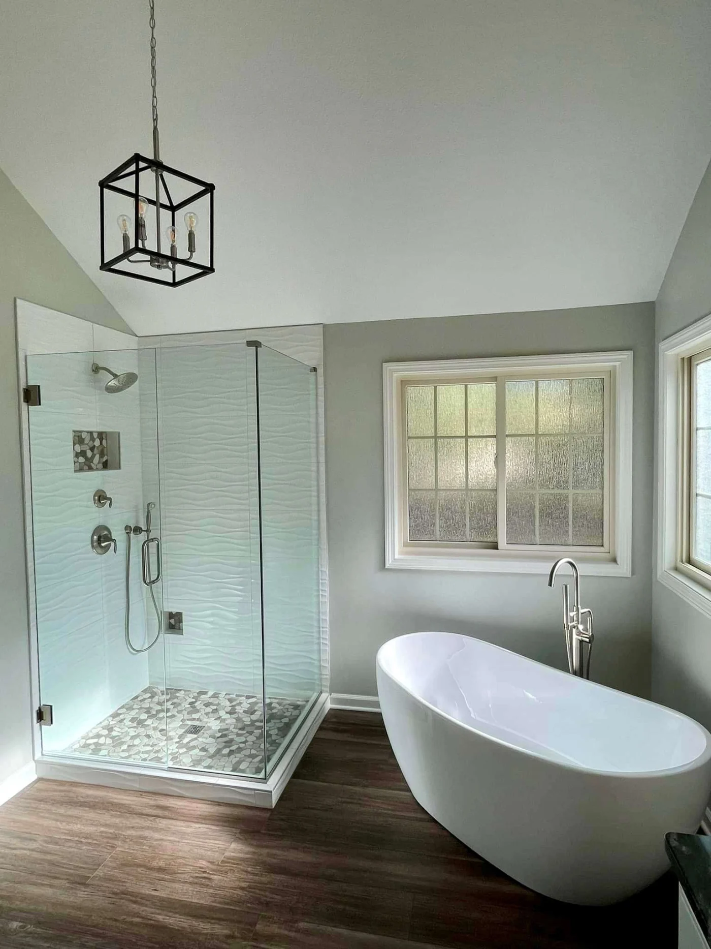 Stunning Bathroom Remodeling In Lenexa, KS | Jericho A gray bathroom with wooden flooring, a white bathtub, a walk-in shower with glass walls, and windows.